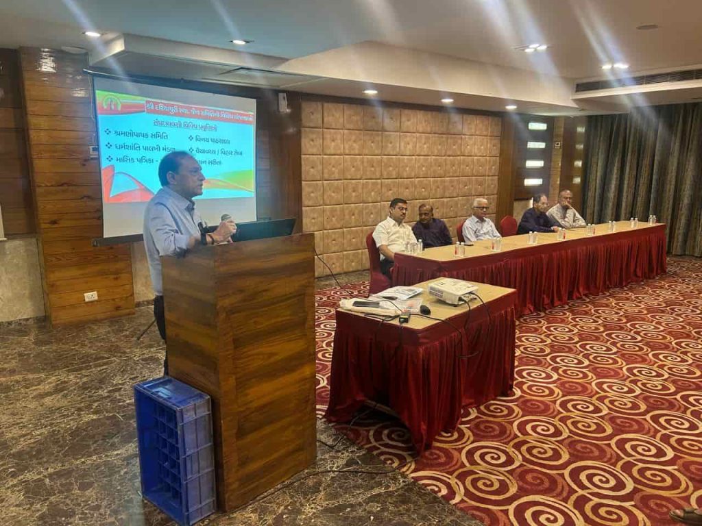 Speaker addressing the audience during a Dariyapuri Sthanakwasi Jain Samiti event in Ahmedabad, with committee members seated on stage and a presentation screen in the background.