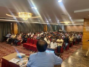 Speaker addressing community members of Dariyapuri Sthanakwasi Jain Samiti seated in a hall with red chairs during a gathering in Ahmedabad.